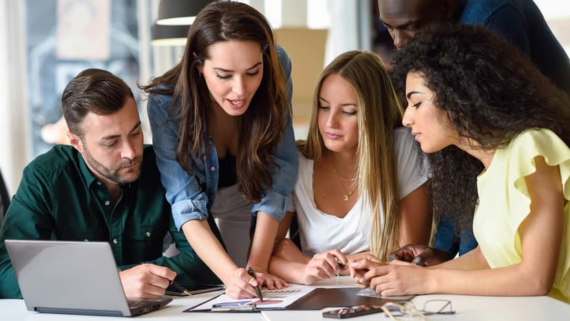 A diverse group of professionals collaborating around a table in a bright office, reviewing charts and using a laptop.